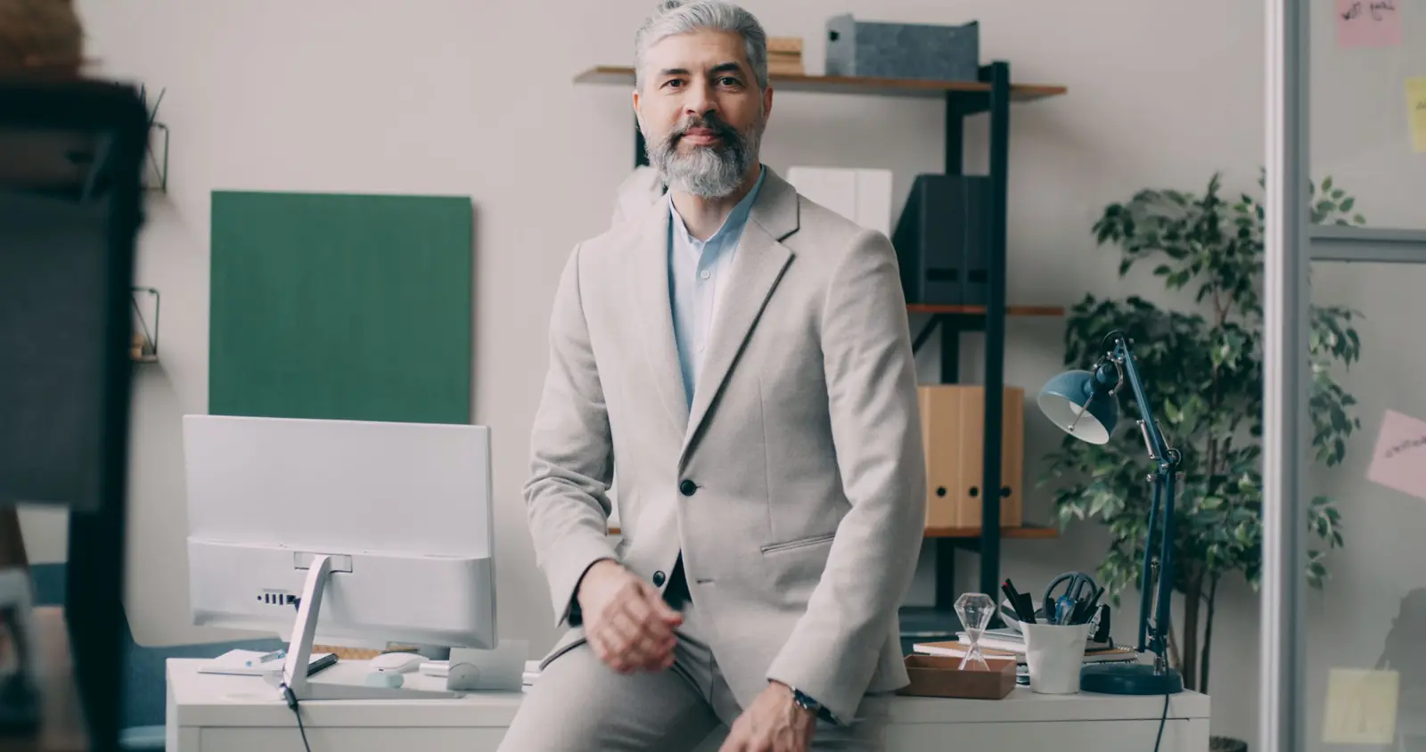 A business owner in a light suit, sitting on the edge of his desk.